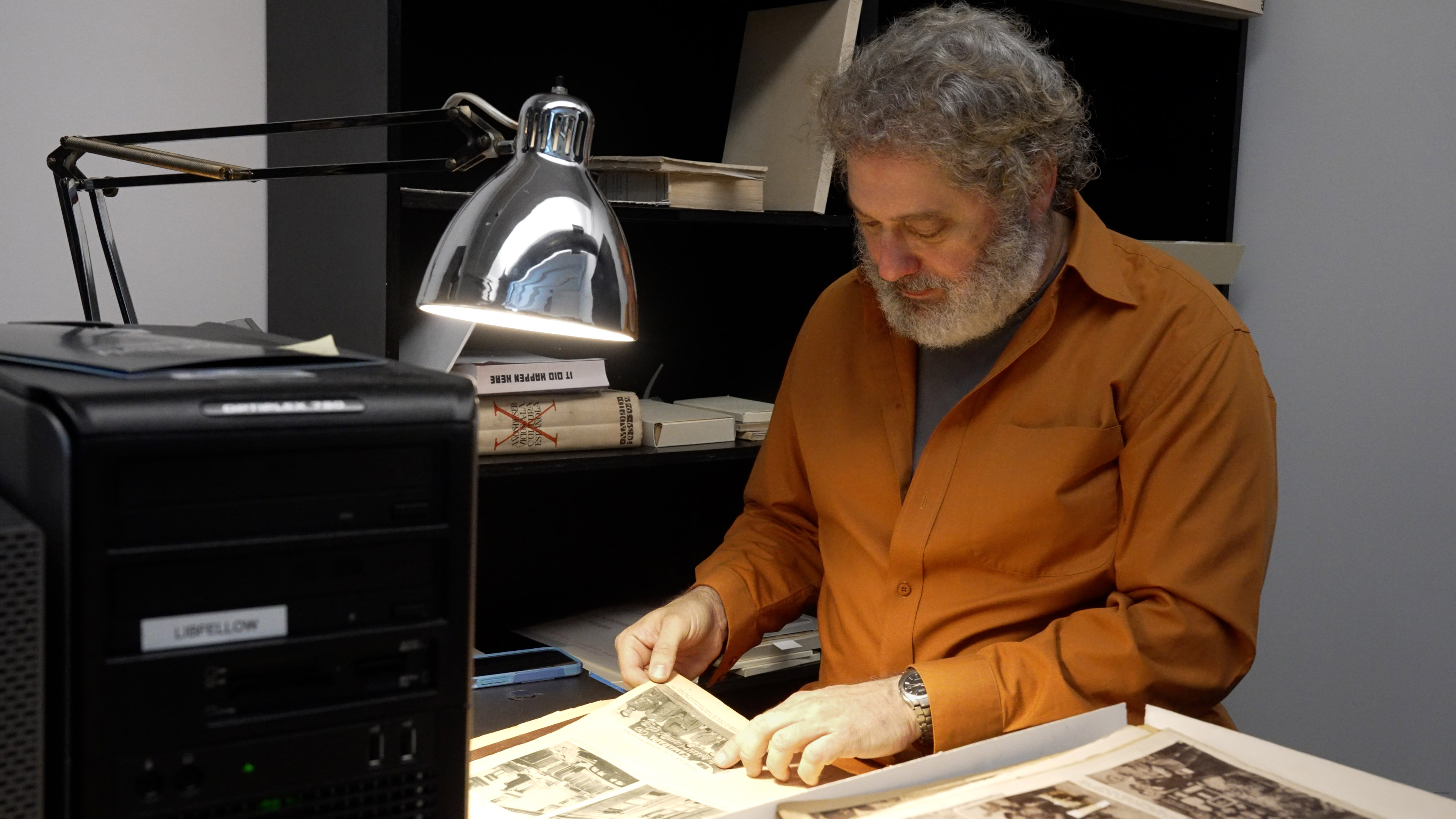 A person with gray hair and a beard, wearing an orange shirt, sits at a desk examining black-and-white photographs under a desk lamp. Stacks of archival materials are visible nearby.