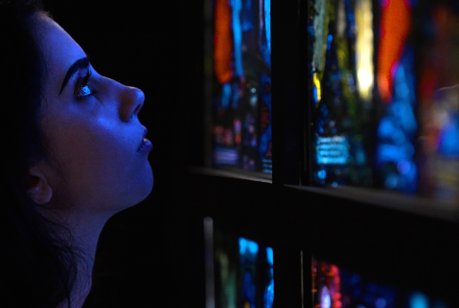 A person with face in profile in a dark gallery looking closely at a stained glass window.